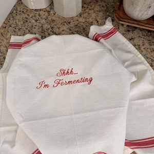Puede incluir: Un pa&ntilde;o de cocina blanco con texto bordado en rojo que dice "Shhh... I'm Fermenting" en una fuente cursiva. El pa&ntilde;o tiene rayas horizontales rojas a lo largo del borde inferior y est&aacute; drapeado sobre un objeto, mostrando una textura tejida.