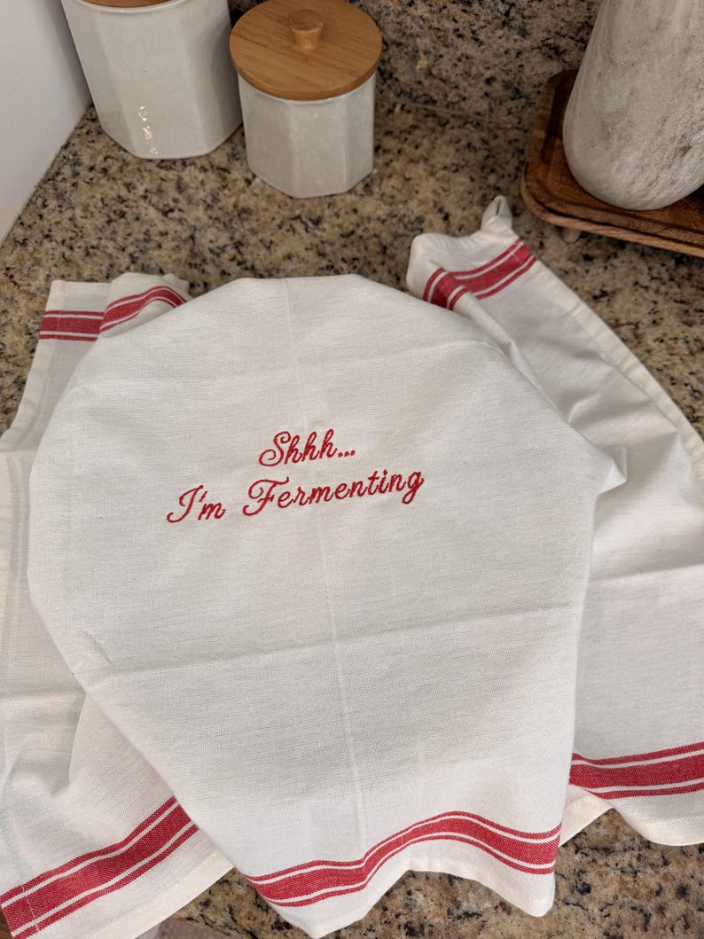 Puede incluir: Un pa&ntilde;o de cocina blanco con texto bordado en rojo que dice "Shhh... I'm Fermenting" en una fuente cursiva. El pa&ntilde;o tiene rayas horizontales rojas a lo largo del borde inferior y est&aacute; drapeado sobre un objeto, mostrando una textura tejida.