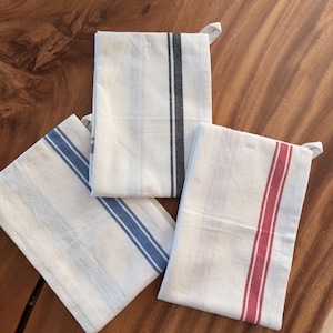Puede incluir: Tres pa&ntilde;os de cocina rectangulares doblados cuidadosamente. Cada pa&ntilde;o es de color blanco roto con un patr&oacute;n de rayas verticales distintivo. Uno tiene una raya negra, otro una raya azul y el tercero muestra una raya de color rojo vivo.