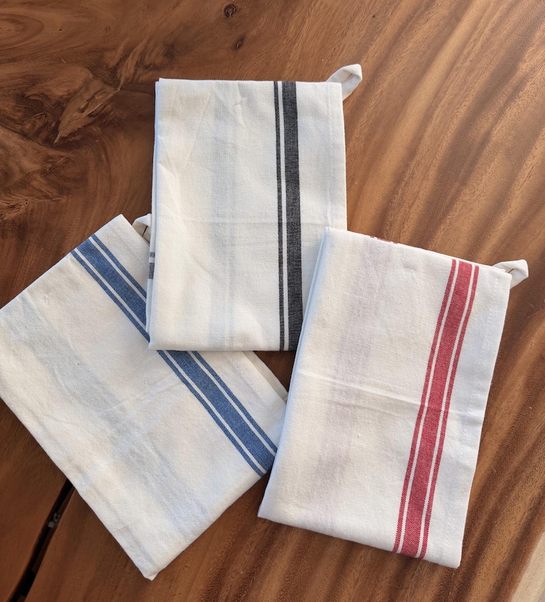 Puede incluir: Tres pa&ntilde;os de cocina rectangulares doblados cuidadosamente. Cada pa&ntilde;o es de color blanco roto con un patr&oacute;n de rayas verticales distintivo. Uno tiene una raya negra, otro una raya azul y el tercero muestra una raya de color rojo vivo.
