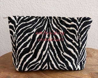 May include: A zippered pouch featuring a black and white zebra stripe pattern. The front of the pouch has the text embroidery essentials stitched in red thread across the center. The pouch has a rectangular shape with a top zipper closure.