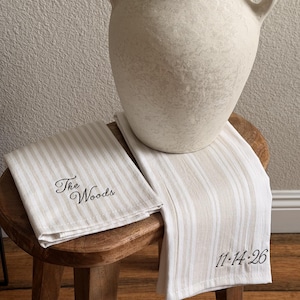 May include: Two folded kitchen towels featuring a beige and white vertical striped pattern. One towel is embroidered with the text "The Woods" in black script, while the other towel features the embroidered date "11-14-26" in black script.