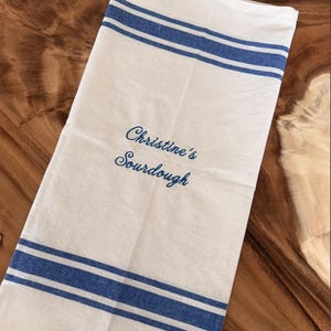 Puede incluir: Un pa&ntilde;o de cocina rectangular de color blanco con dos franjas horizontales azules en los bordes superior e inferior. El centro de la tela tiene bordado el texto azul "Christine's Sourdough" en una fuente de escritura cursiva.