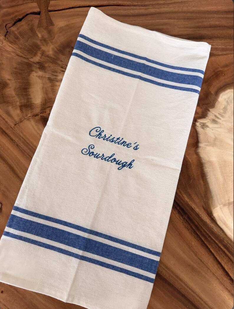 Puede incluir: Un pa&ntilde;o de cocina rectangular de color blanco con dos franjas horizontales azules en los bordes superior e inferior. El centro de la tela tiene bordado el texto azul "Christine's Sourdough" en una fuente de escritura cursiva.