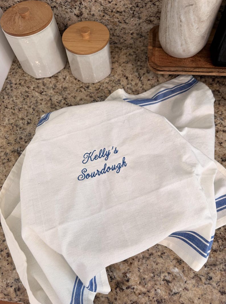 Puede incluir: Un pa&ntilde;o de cocina blanco con rayas azules y el texto bordado Kelly's Sourdough en letra cursiva azul. El pa&ntilde;o est&aacute; colocado sobre un objeto redondo, mostrando una textura de tela tejida y bordes limpios, permitiendo ver claramente el bordado.