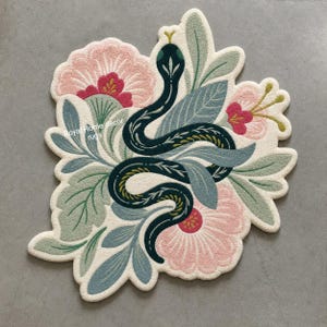 Può includere: Tappeto decorativo con un motivo a serpente stilizzato, intrecciato con fiori rosa e foglie verdi e blu. Il tappeto ha uno sfondo color crema e il testo "Royal Home decor rugs".
