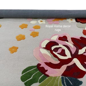 May include: A floral area rug with a light gray background and a large rose design in shades of pink, red, and green. Scattered around the rose are smaller orange and white flower accents. The text "Royal Home decor rugs" is visible.