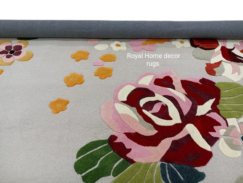 May include: A floral area rug with a light gray background and a large rose design in shades of pink, red, and green. Scattered around the rose are smaller orange and white flower accents. The text "Royal Home decor rugs" is visible.
