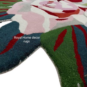 May include: A close-up of a floral rug with a light gray background. The rug features large, stylized flowers in shades of pink, red, white, and green. The text "Royal Home decor rugs" is visible on the rug.