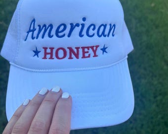 May include: White trucker hat with the text "American HONEY" in blue and red. The hat has a white mesh back and a curved brim. The hat is being held up by a hand with a ring on the ring finger.