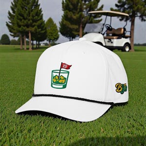 Op de afbeelding: Witte baseballcap met een zwarte touwrand. De cap heeft een geborduurd ontwerp van een cocktail met een golfflag en het nummer 19. De cap heeft ook de letters "By" in een groen en geel ontwerp.