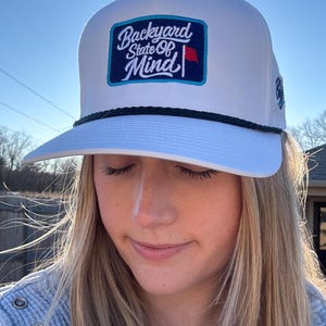 May include: White trucker hat with a curved brim and a black rope detail. The hat features a rectangular patch with the words "Backyard State Of Mind" in a blue and white design, with a small red flag.