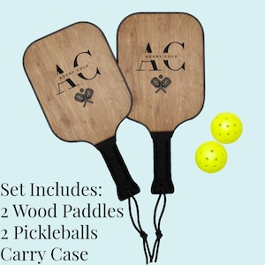 Könnte beinhalten: Ein Pickleball-Set mit zwei Holzschlägern mit schwarzen Griffen und dem Text "AC AVERY COLE". Zwei gelbe Picklebälle sind ebenfalls enthalten. Für Pickleball-Enthusiasten.
