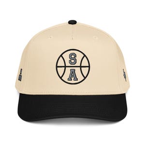 May include: A beige baseball cap with a black brim and a black button on top. The front features an embroidered black basketball graphic with the letters S and A in grey. Small embroidered icons are visible on the left and right side panels.