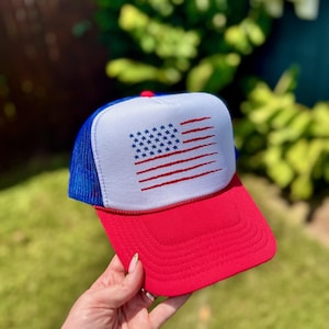 May include: A red, white, and blue trucker hat with an American flag design. The hat has a red peak, a white front panel with a flag design, and a blue mesh back. The flag design features blue stars and red stripes.