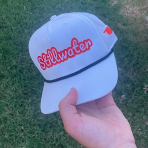 Könnte beinhalten: Weiße Baseballkappe mit dem Wort "Stillwater" in roter Schrift gestickt. Die Kappe hat ein schwarzes Seildetail und einen kleinen orangefarbenen Akzent an der Seite. Der Hut wird in einer Hand vor einem Hintergrund aus grünem Gras gehalten.