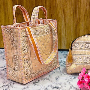 May include: A set of two beige and pink patterned bags. One is a tote bag with orange straps, and the other is a small, rounded cosmetic bag with a gold tassel. A bouquet of pink and white flowers sits in front of the bags.