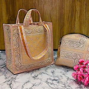 May include: A matching set of beige tote bag and cosmetic bag with a paisley design. The tote bag has two handles and a shoulder strap. The cosmetic bag has a zipper closure. Pink flowers are in the foreground.