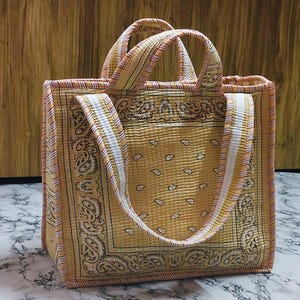 May include: A quilted tote bag with a yellow paisley pattern. The bag has a rectangular shape with matching handles and a white and yellow striped strap. The bag is made of a soft, quilted fabric.
