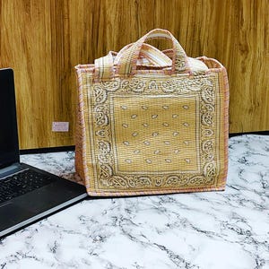 May include: A square tote bag with a paisley pattern in shades of yellow and tan. The bag has a matching handle and trim, and is sitting on a marble surface. The bag is next to a laptop.