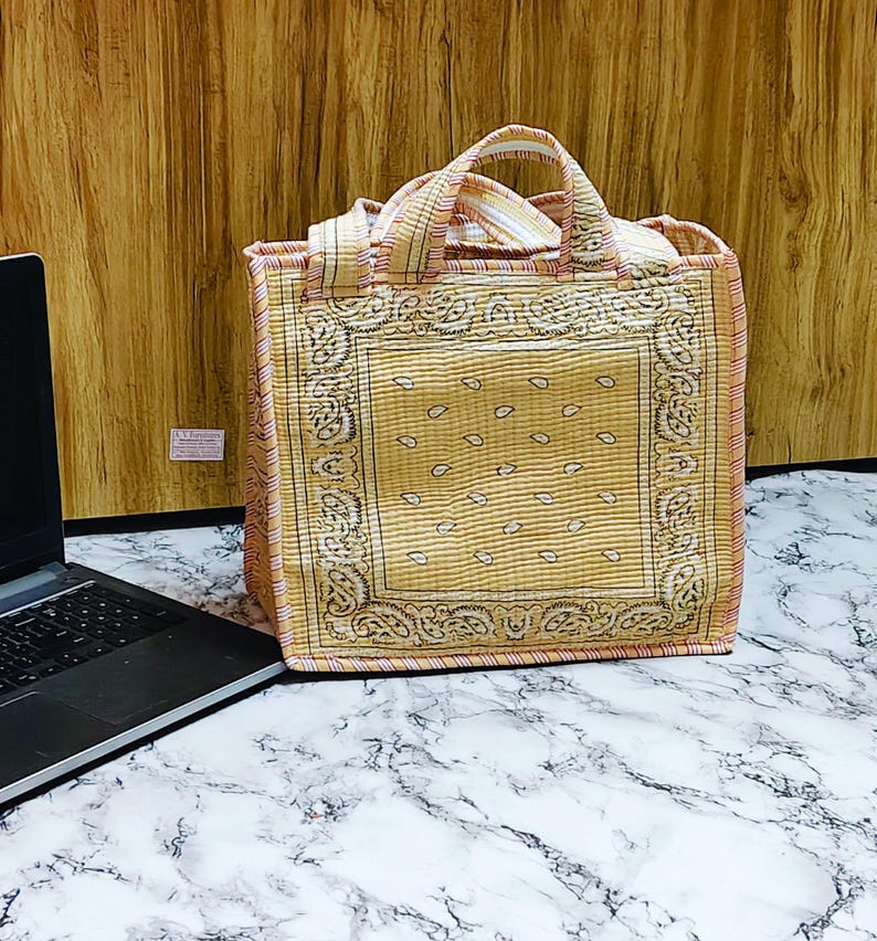 May include: A square tote bag with a paisley pattern in shades of yellow and tan. The bag has a matching handle and trim, and is sitting on a marble surface. The bag is next to a laptop.