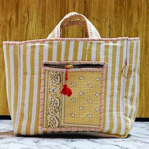 May include: A quilted tote bag with vertical yellow and white stripes. It features a front pocket with a paisley design and a zipper closure. A red tassel hangs from the zipper. The bag has two handles and a gold-colored ring attached to the side.