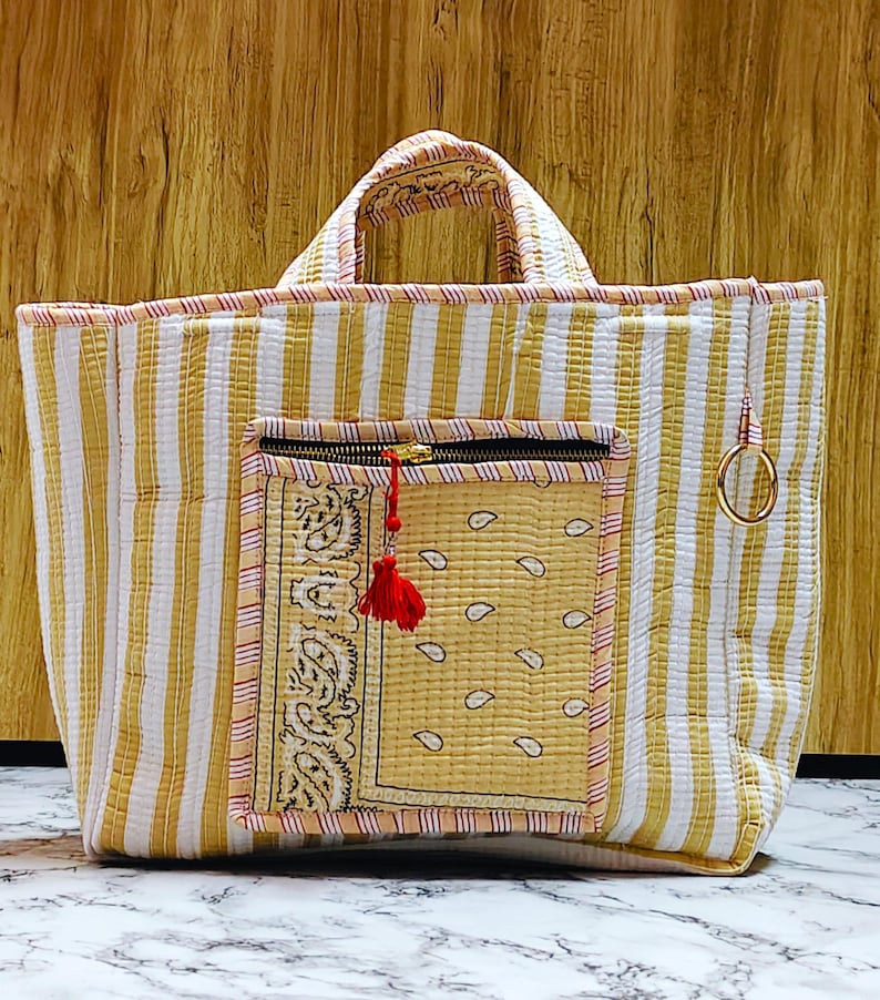 May include: A quilted tote bag with vertical yellow and white stripes. It features a front pocket with a paisley design and a zipper closure. A red tassel hangs from the zipper. The bag has two handles and a gold-colored ring attached to the side.