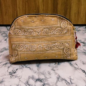 May include: A tan, quilted cosmetic bag with a black zipper and a paisley pattern. The bag has a rounded shape and a small red tassel attached to the zipper pull. The bag is sitting on a marble surface.