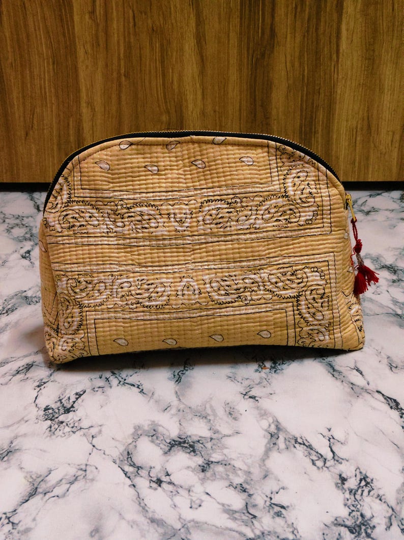 May include: A tan, quilted cosmetic bag with a black zipper and a paisley pattern. The bag has a rounded shape and a small red tassel attached to the zipper pull. The bag is sitting on a marble surface.