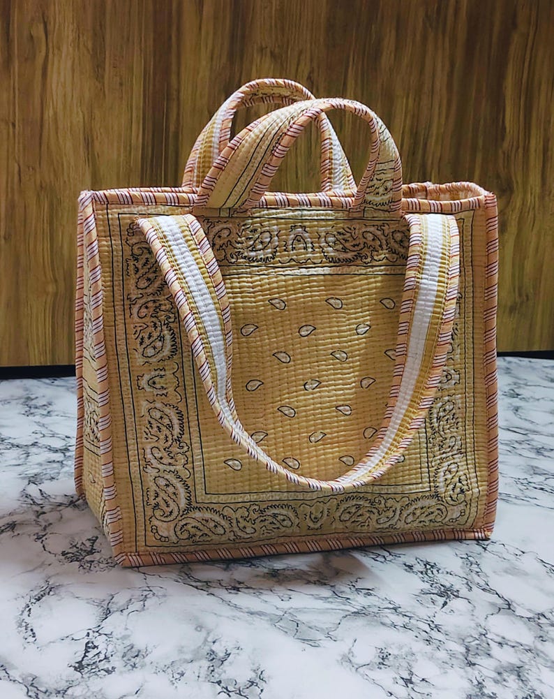 May include: A quilted tote bag with a yellow paisley pattern. The bag has a rectangular shape with matching handles and a white and yellow striped strap. The bag is made of a soft, quilted fabric.