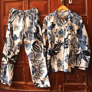 May include: A two-piece pajama set with a botanical print. The set includes a button-down shirt and drawstring pants. The fabric is white with a pattern of blue and teal leaves, flowers, and birds. The pajamas are hanging on a wooden door.
