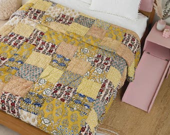 May include: A patchwork quilt featuring a variety of yellow, white, and blue floral and geometric patterns. The quilt is draped over a bed with a pink upholstered headboard and two white pillows with pom-pom trim edges.