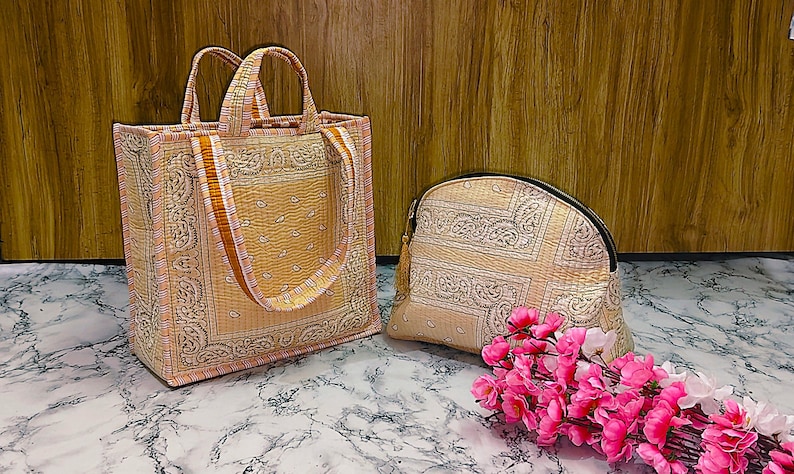 May include: A matching set of beige tote bag and cosmetic bag with a paisley design. The tote bag has two handles and a shoulder strap. The cosmetic bag has a zipper closure. Pink flowers are in the foreground.
