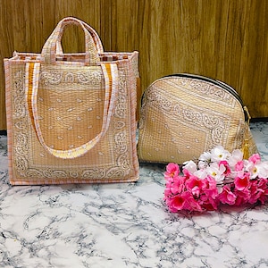 May include: Two-piece set of paisley-patterned bags in beige and peach tones. The set includes a tote bag with handles and a zippered pouch. A bundle of pink and white flowers rests in front of the bags.