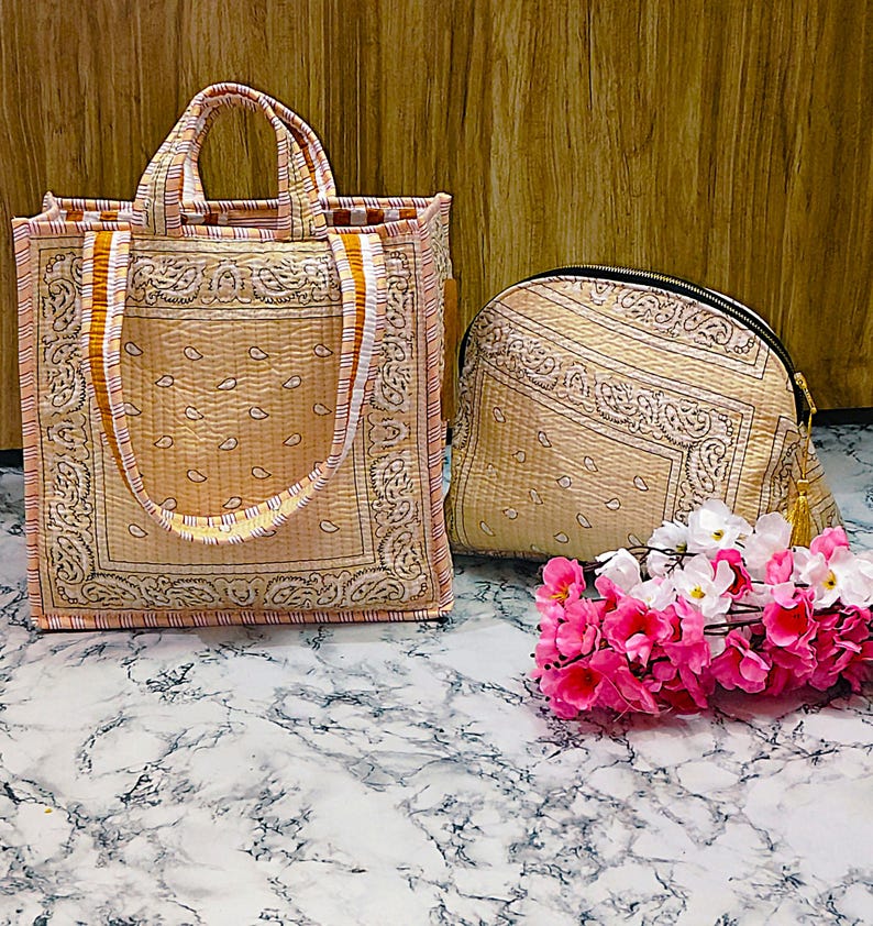 May include: Two-piece set of paisley-patterned bags in beige and peach tones. The set includes a tote bag with handles and a zippered pouch. A bundle of pink and white flowers rests in front of the bags.