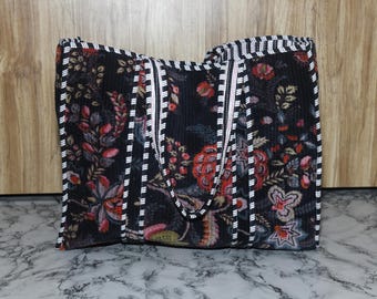May include: A rectangular quilted tote bag featuring a dark black base with a multi-colored floral pattern in shades of red, pink, and green. The bag includes two shoulder straps and edges trimmed with a black and white striped fabric border.