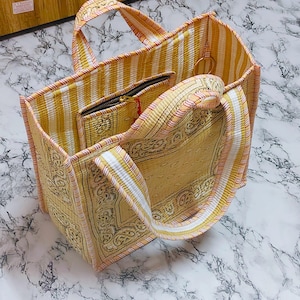 May include: A yellow and white striped tote bag with matching handles and trim. The bag features a patterned design on the front and sides, and includes a small zippered pouch inside. The bag is open, revealing the interior.