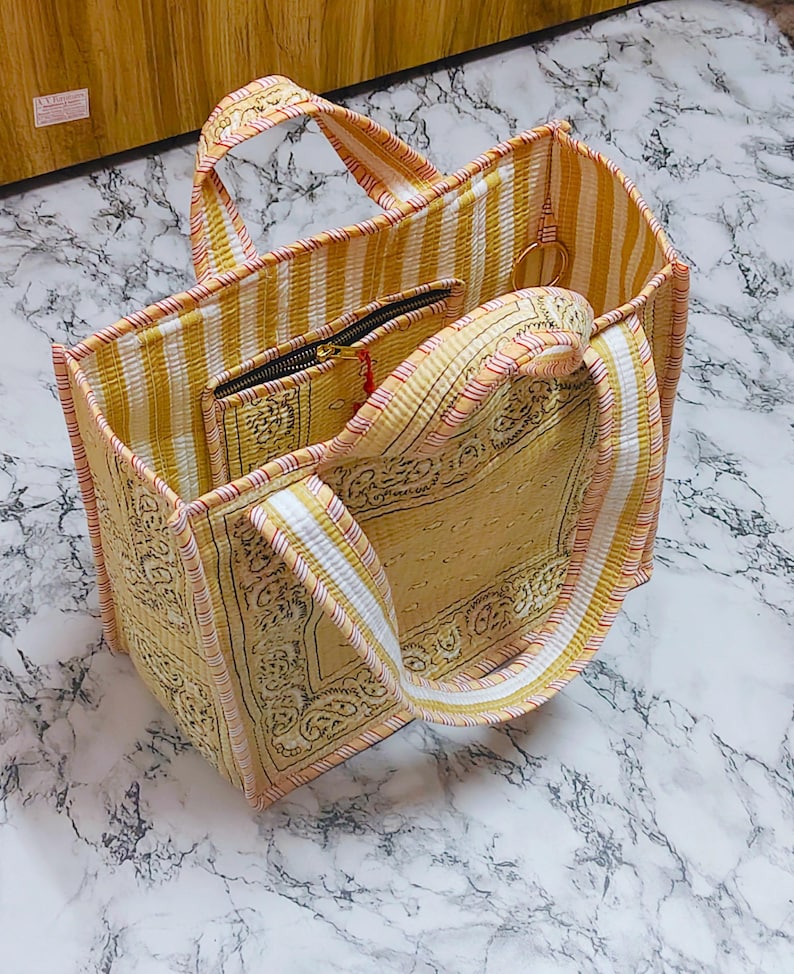 May include: A yellow and white striped tote bag with matching handles and trim. The bag features a patterned design on the front and sides, and includes a small zippered pouch inside. The bag is open, revealing the interior.