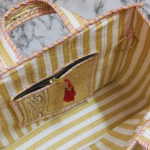 May include: Open view of a striped tote bag with a zippered pocket. The bag features yellow and white vertical stripes, a black zipper, and a red tassel. The bag's trim is a light peach color with red accents. A gold ring is attached to the bag.