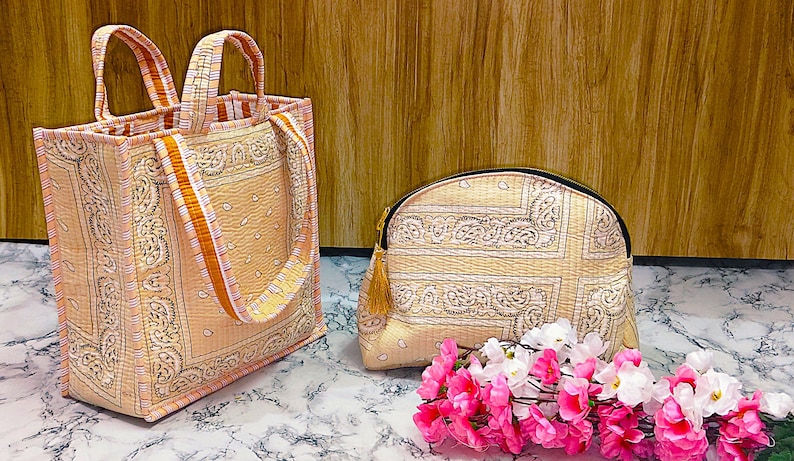 May include: A set of two beige and pink patterned bags. One is a tote bag with orange straps, and the other is a small, rounded cosmetic bag with a gold tassel. A bouquet of pink and white flowers sits in front of the bags.