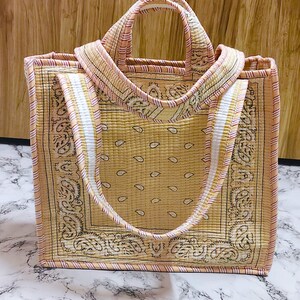 May include: A quilted tote bag with a paisley pattern in shades of yellow and black. The bag features a rectangular shape with sturdy handles and a striped trim in pink and white. The bag is designed for carrying items.