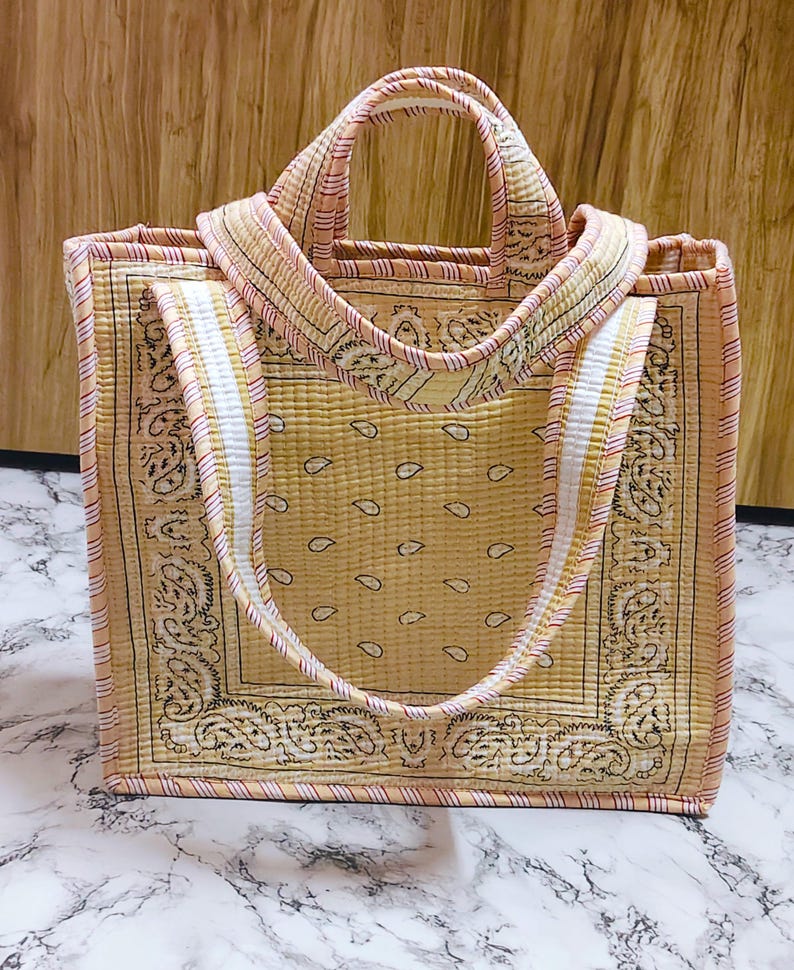 May include: A quilted tote bag with a paisley pattern in shades of yellow and black. The bag features a rectangular shape with sturdy handles and a striped trim in pink and white. The bag is designed for carrying items.