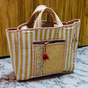 May include: A handmade tote bag with a striped pattern in yellow and white. The bag features a front pocket with a paisley design and a red tassel zipper pull. The handles are also striped, and a gold ring is attached to the side.