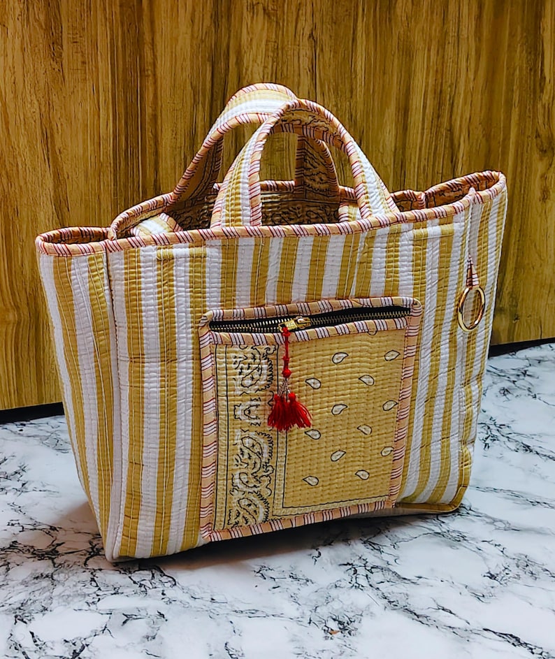 May include: A handmade tote bag with a striped pattern in yellow and white. The bag features a front pocket with a paisley design and a red tassel zipper pull. The handles are also striped, and a gold ring is attached to the side.