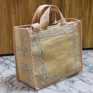 May include: A quilted tote bag with a yellow paisley pattern. The bag has two handles and is trimmed with a pink and white striped fabric. The bag is rectangular and appears to be made of fabric.