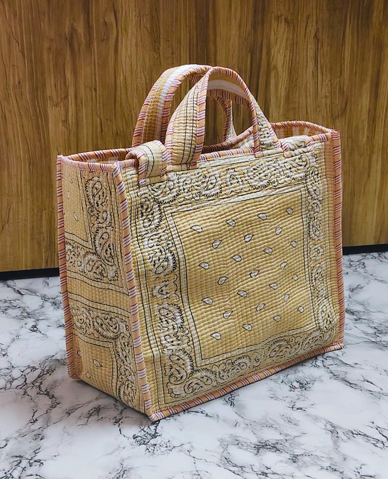 May include: A quilted tote bag with a yellow paisley pattern. The bag has two handles and is trimmed with a pink and white striped fabric. The bag is rectangular and appears to be made of fabric.