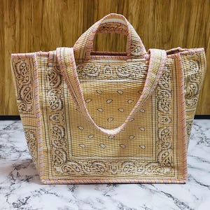 May include: A quilted tote bag with a bandana print in shades of tan, cream, and pink. The bag features a rectangular shape with sturdy handles and a matching strap. The design includes paisley patterns and a striped border.