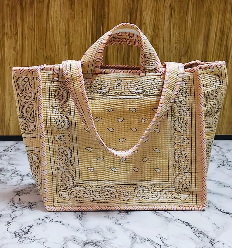 May include: A quilted tote bag with a bandana print in shades of tan, cream, and pink. The bag features a rectangular shape with sturdy handles and a matching strap. The design includes paisley patterns and a striped border.