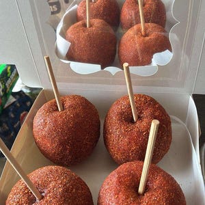 May include: Six red candy apples coated in a chili powder mixture, each with a wooden stick inserted. The apples are arranged in a clear plastic box with a decorative cutout on the lid, perfect for a sweet treat.