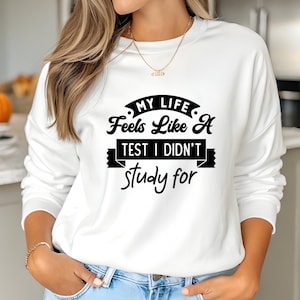 May include: White sweatshirt with the text "MY LIFE Feels Like A TEST I DIDN'T study for" in black. The sweatshirt has long sleeves and a crew neck. A gold necklace is visible.